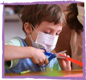 a little boy using safety scissors on construction paper while wearing a face mask